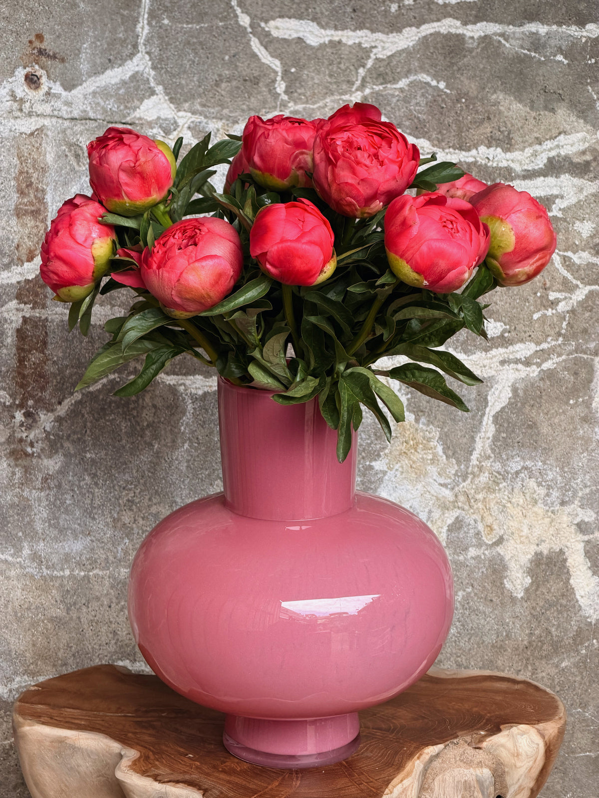 Roze glazen vaas met bolle buik, gevuld met koraalroze pioenen, op een houten boomstamtafel