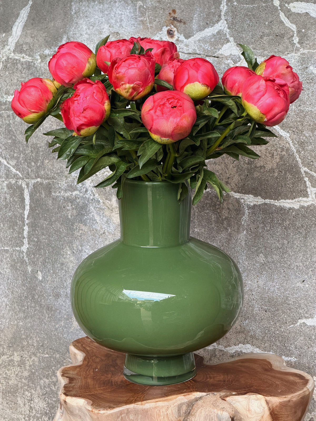Bolle olijfgroene glazen vaas met korte hals, gevuld met koraalroze pioenen, op een houten boomstamtafel