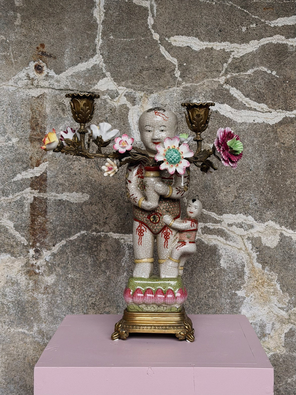 Porseleinen kandelaar met kindfiguur en bronzen bloemtakken op een roze sokkel tegen een betonnen muur