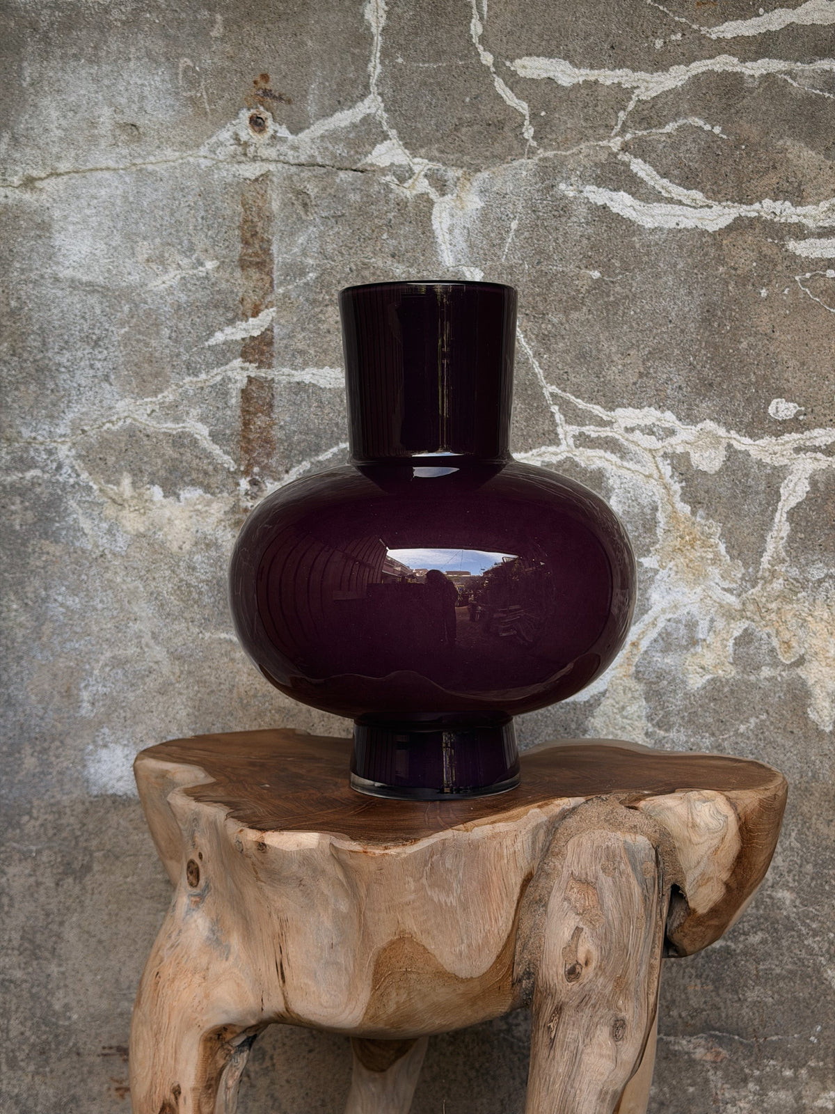 Lege aubergine glasvaas met brede bolle buik, rechte hals en voet op een houten boomstamkruk