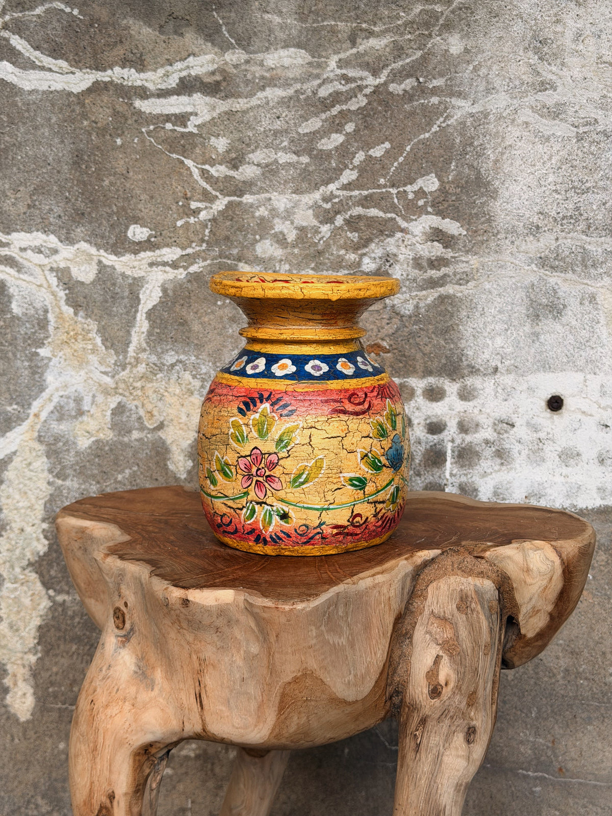 Kleine gele melkpot met bloemdecor op een boomstamkruk tegen een ruwe betonnen muur