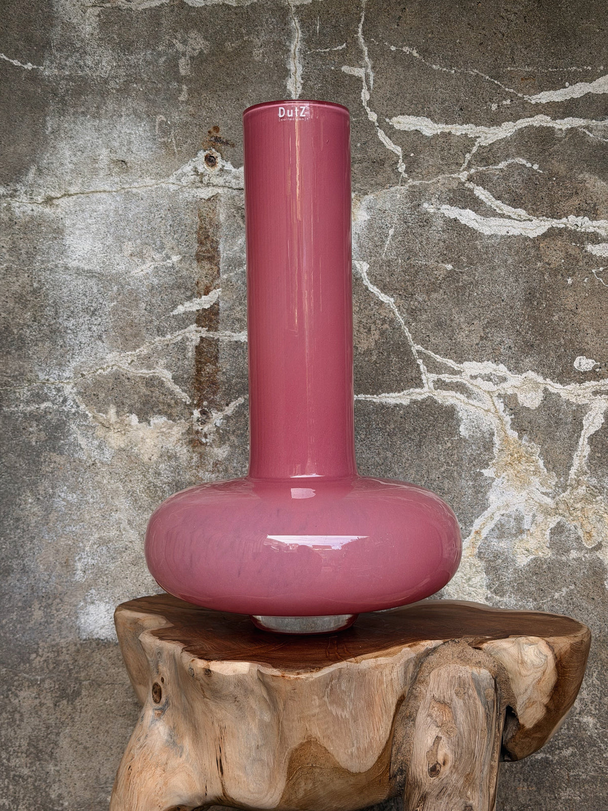 Roze glazen vaas met platte bolle voet en lange smalle hals met DutZ-logo, op een houten kruk