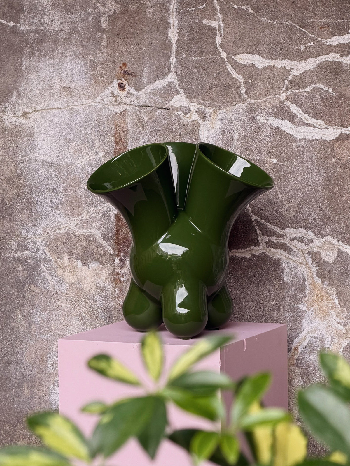 De groene vaas van een afstand op de roze sokkel, met bladeren van een plant onscherp op de voorgrond