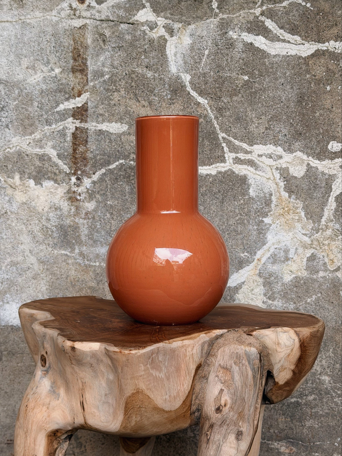 Terracotta-oranje glazen vaas met bolle buik en hoge rechte hals op een houten kruk