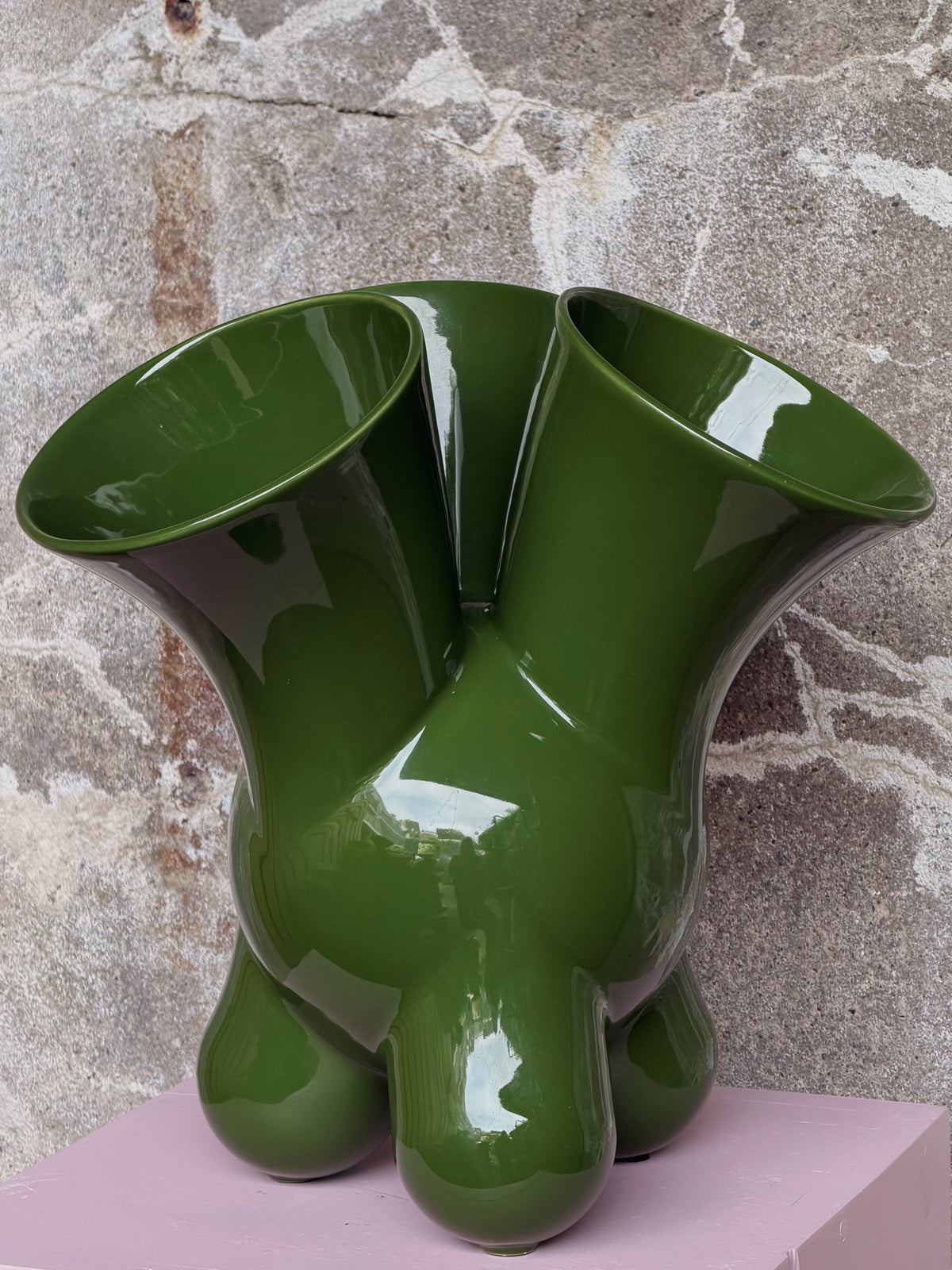 De groene vaas van dichtbij met twee brede openingen en ronde voetjes op de rand van de roze sokkel